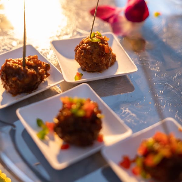 Four Indian meatballs, garnished with chopped vegetables, served on small white square plates.