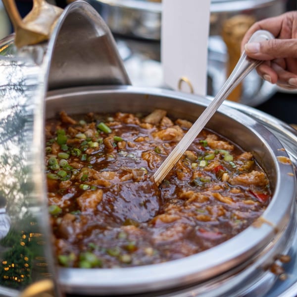 A hand serving Indian chili chicken from a silver chafing dish, garnished with green onions.