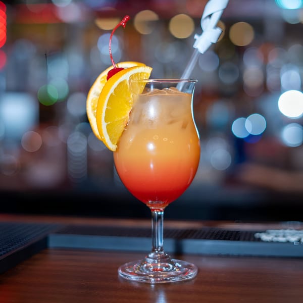 A vibrant orange and red cocktail, garnished with orange slices and a cherry, sits on a bar.