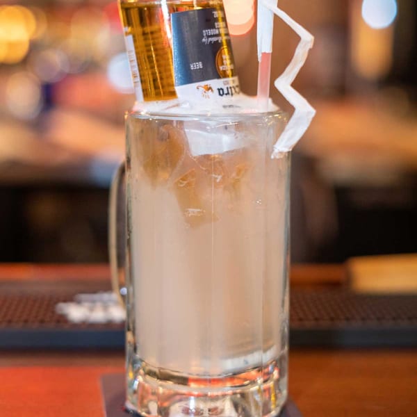 A beer bottle inverted into a mug of light-colored drink with ice and a straw.