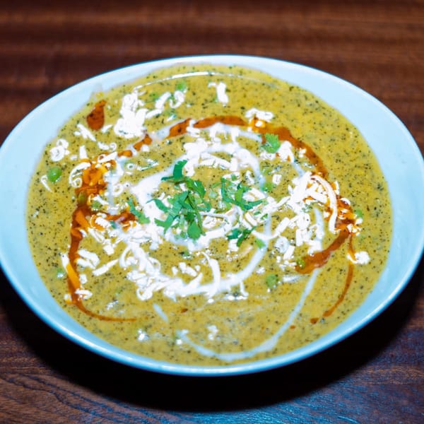 A bowl of Indian curry, garnished with white cream, red oil, and fresh green herbs.