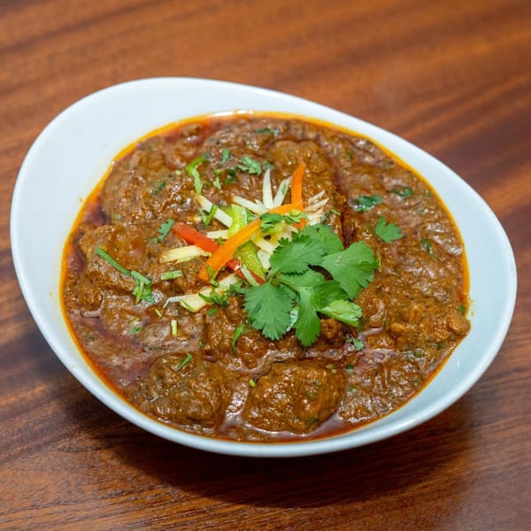 Rich brown Indian curry with meat, garnished with fresh cilantro and julienned vegetables in a white bowl.