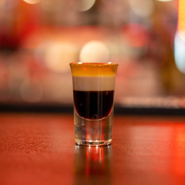 A layered shot glass with dark, white, and yellow liquids, reflecting on a red surface.