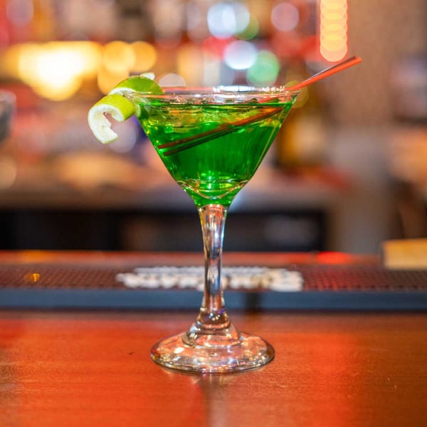 Vibrant green cocktail in a martini glass, garnished with a lime twist and red straw, on a bar.