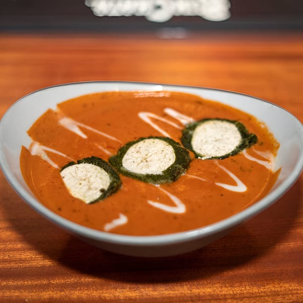A bowl of orange Indian curry with white cream swirls and three paneer discs on a wooden table.