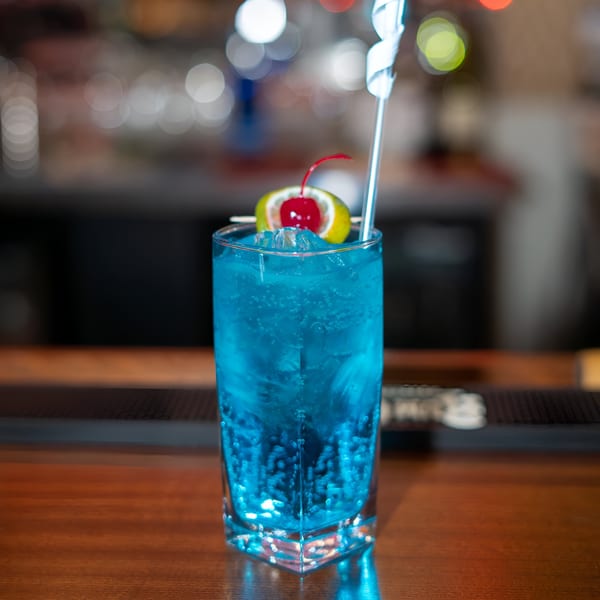 Blue cocktail with a lime and cherry garnish, and a swizzle stick, on a wooden bar.