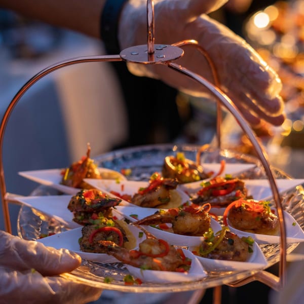 Gloved hands holding a tiered stand with Indian appetizers, garnished with peppers and herbs.