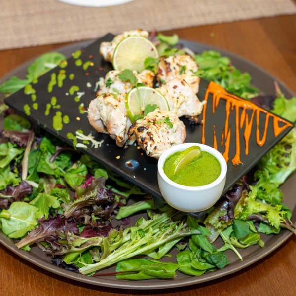 Indian malai tikka chicken with lime, green chutney, and a side salad on a black plate.