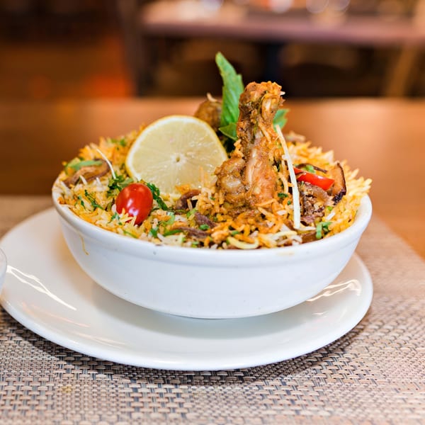 A bowl of Indian chicken biryani with rice, lemon, tomato, and herbs on a woven placemat.