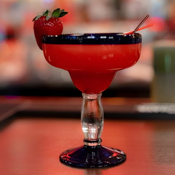 A vibrant red margarita with a strawberry garnish and two straws, on a red surface.